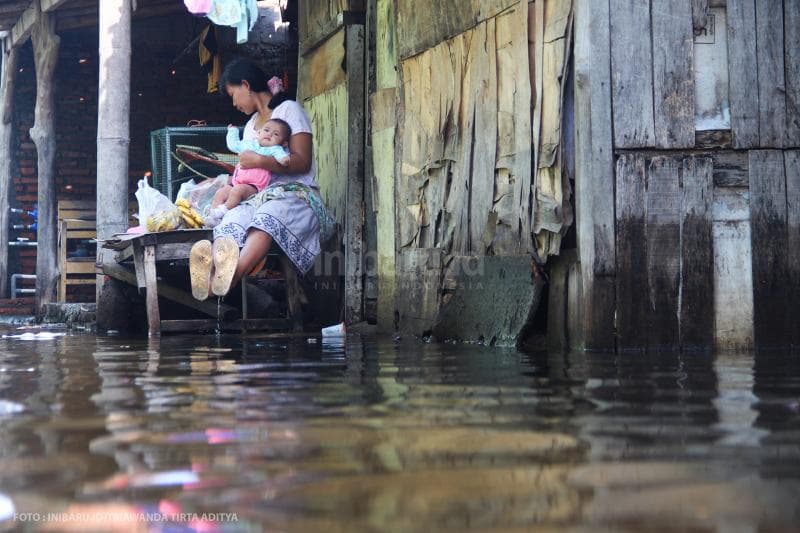 Warga Tambaklorok menggendong bayinya di depan rumah yang tergenang air rob atau banjir rob.<br>